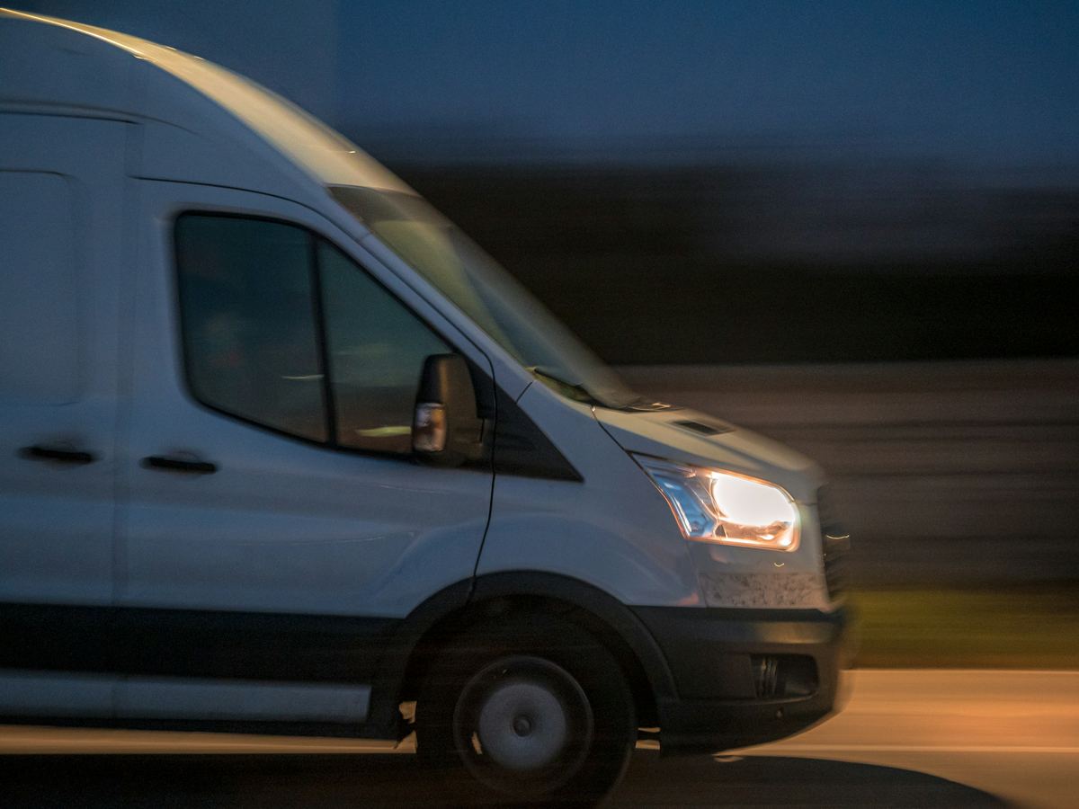 Een witte bestelwagen rijdt met brandende koplampen over de snelweg, bij valavond